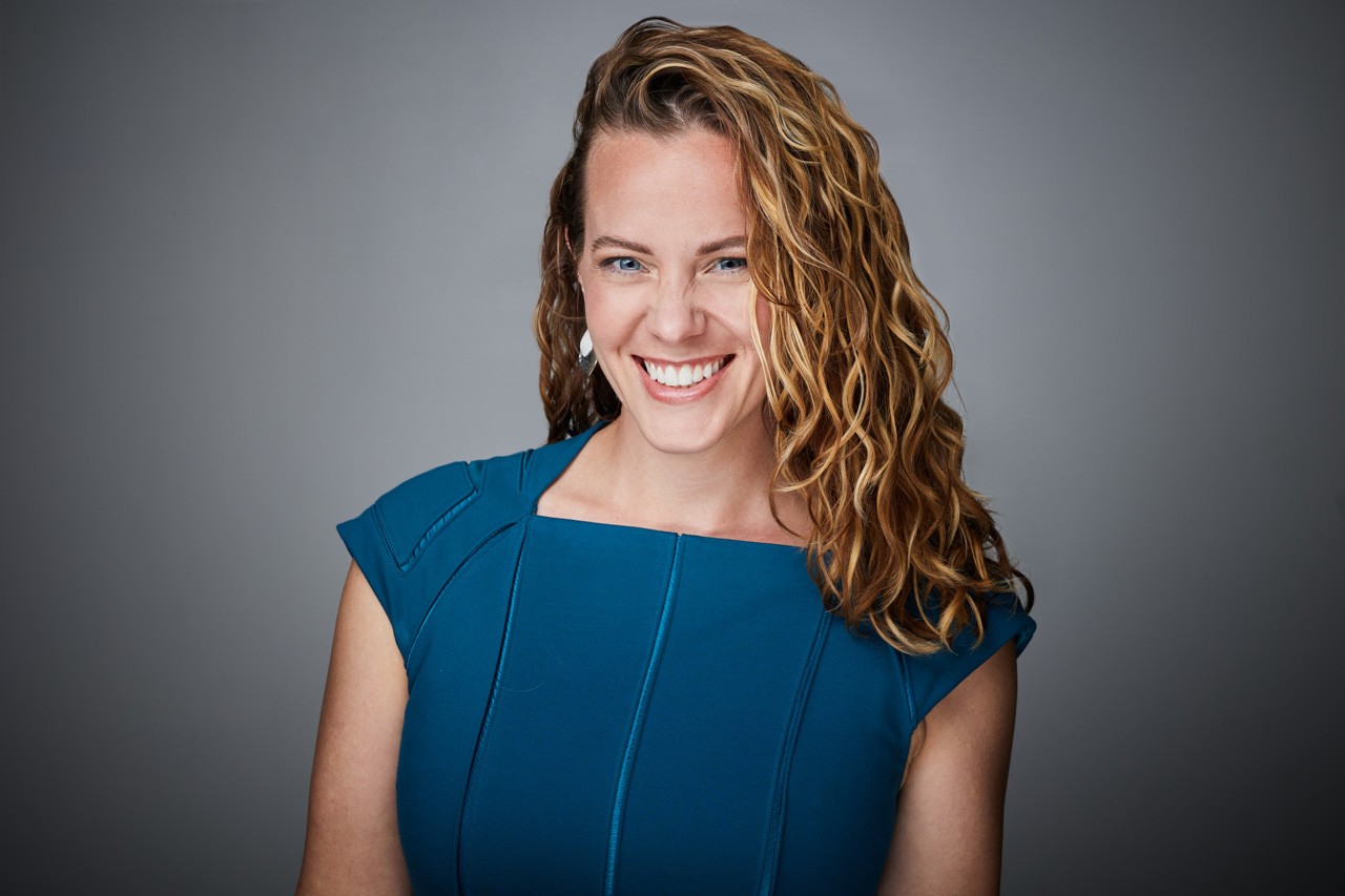 Mindy Herb, smiling, in a teal dress against a grey studio backdrop.