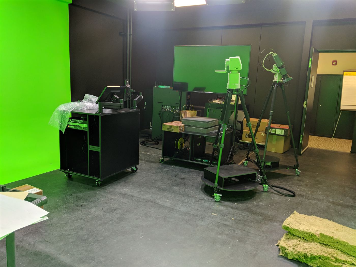 A studio under build, with a lit green screen, equipment racks and robotic camera heads on tripods.
